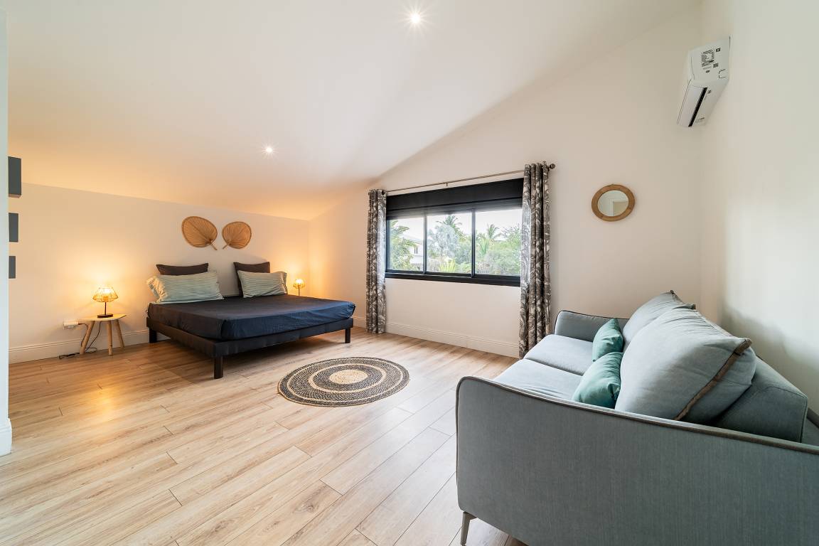 Grande chambre avec lit double, canapé vert, décoration naturelle et fenêtre sur la végétation
