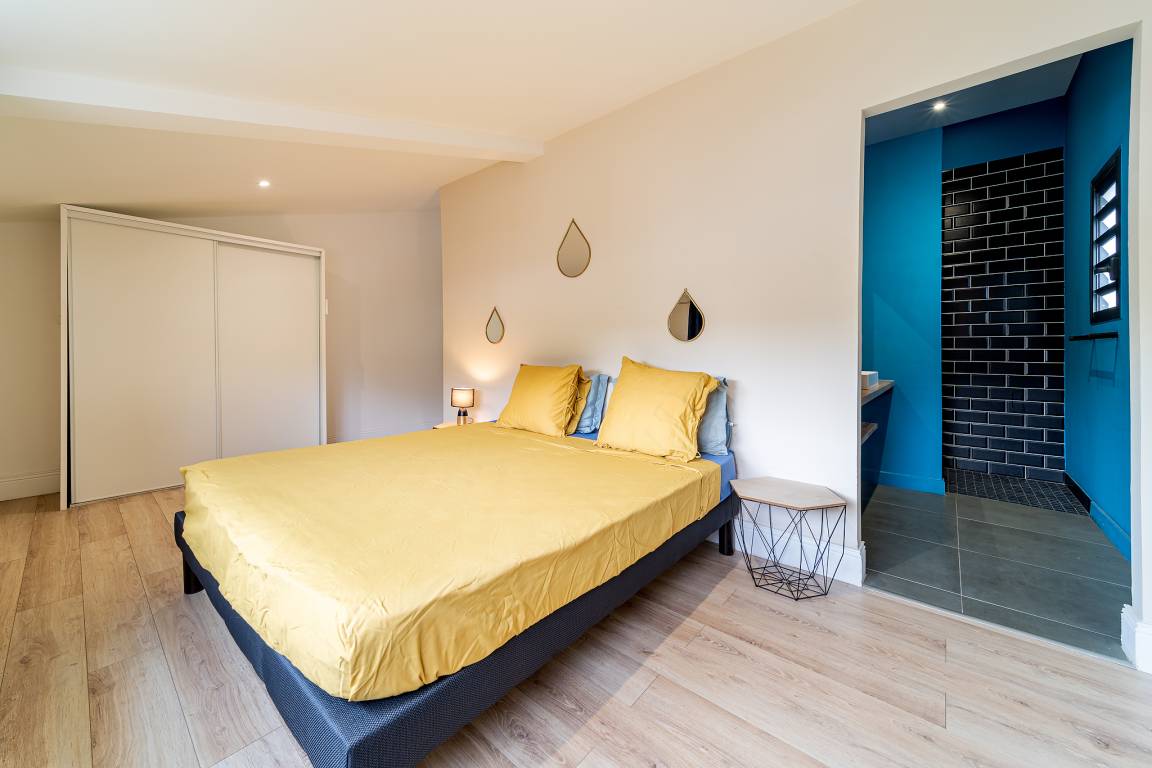 Chambre avec lit double aux draps jaunes et salle d’eau ouverte aux tons bleus